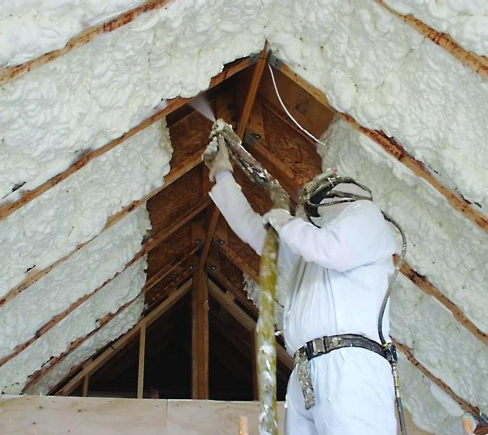 Pracownik w kombinezonie ochronnym aplikuje piankę izolacyjną na drewnianą konstrukcję poddasza, widoczna gęsta warstwa izolacji między krokwiami.